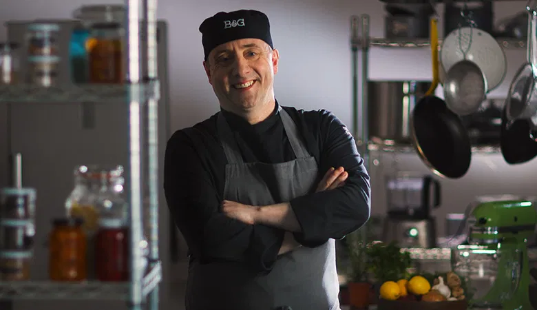 Baxter & Greene chef standing in a kitchen with arms folded, wearing a chef's uniform and apron.